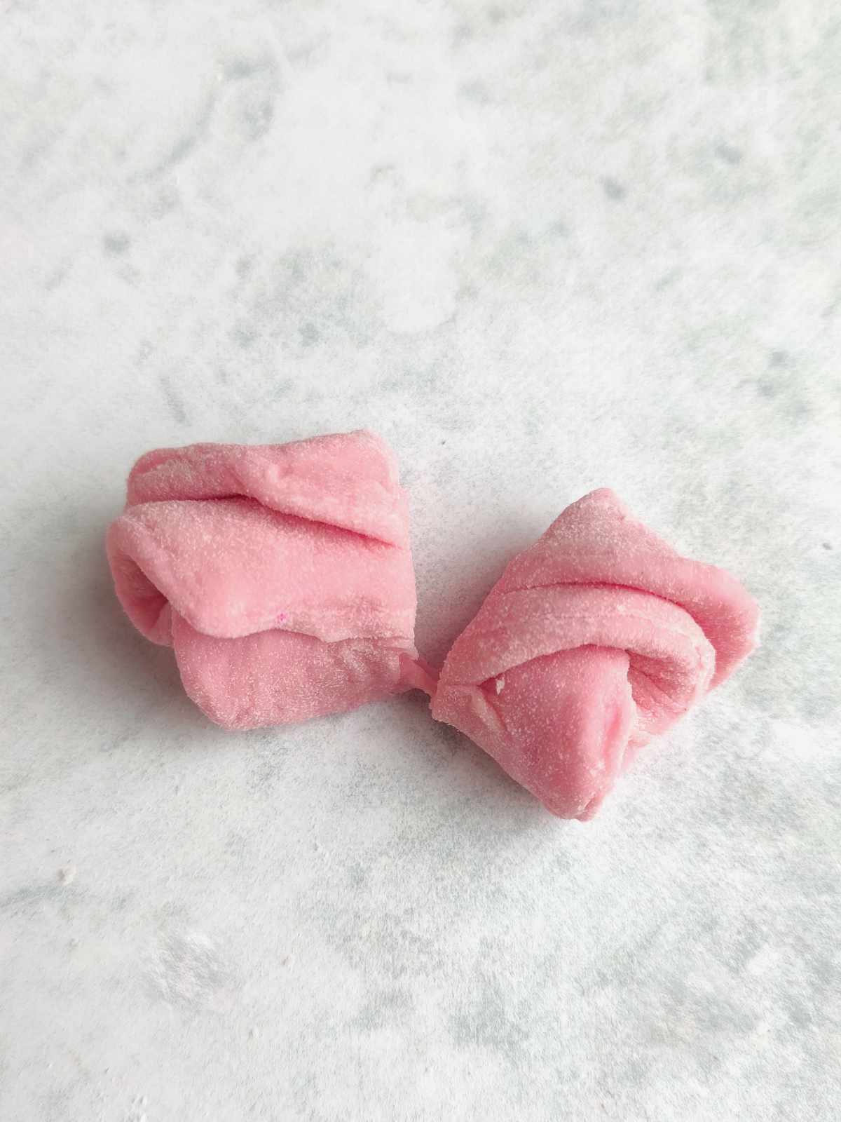 Two pieces of pink, twisted mantou buns sit on a light, textured surface. The treats have a soft, powdery appearance and are shaped with folded, twisted edges.