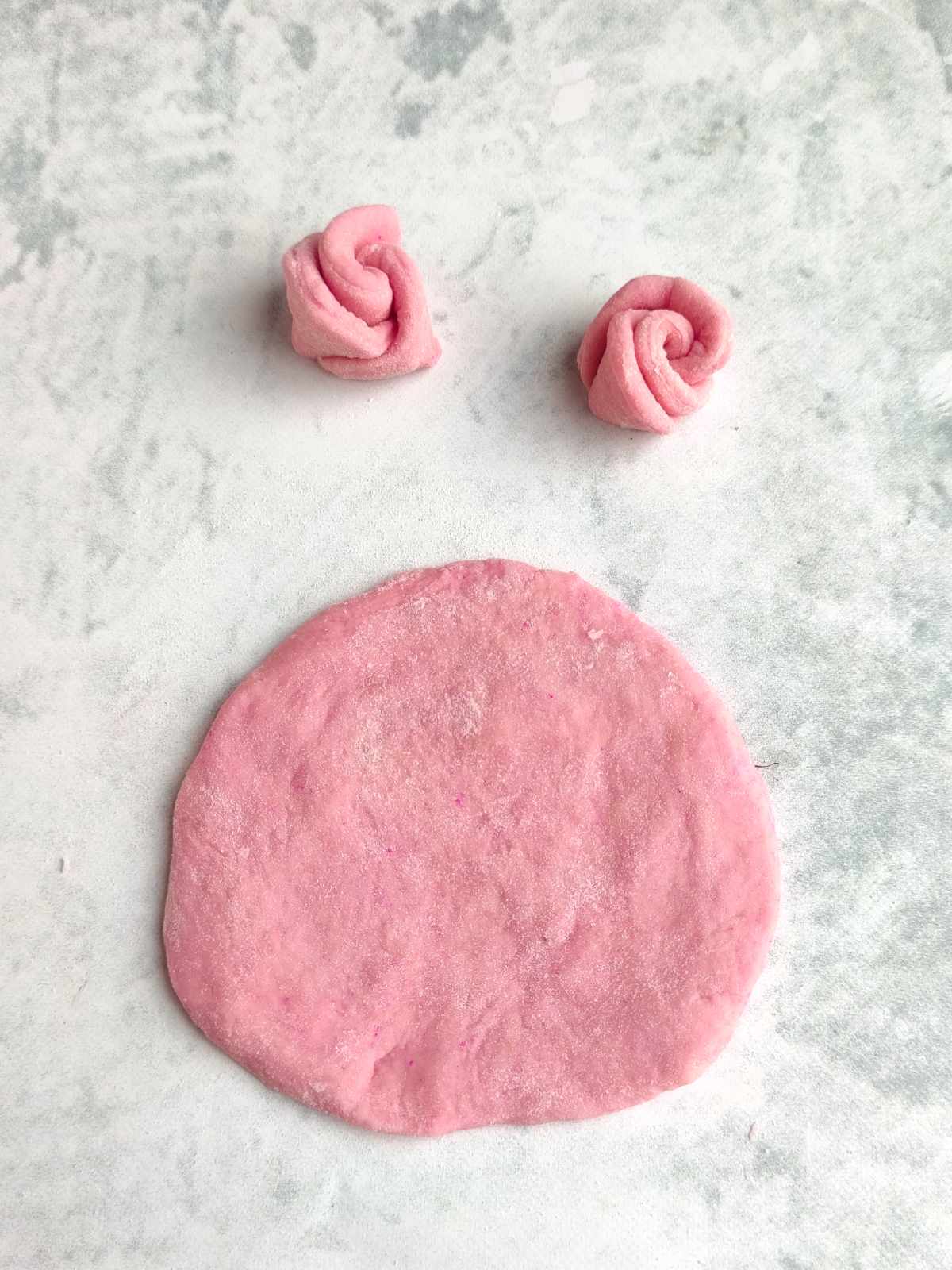 A flat, round piece of pink dough lies on a light surface, with two small, rose-shaped mantou buns made from the same pink dough placed above it.