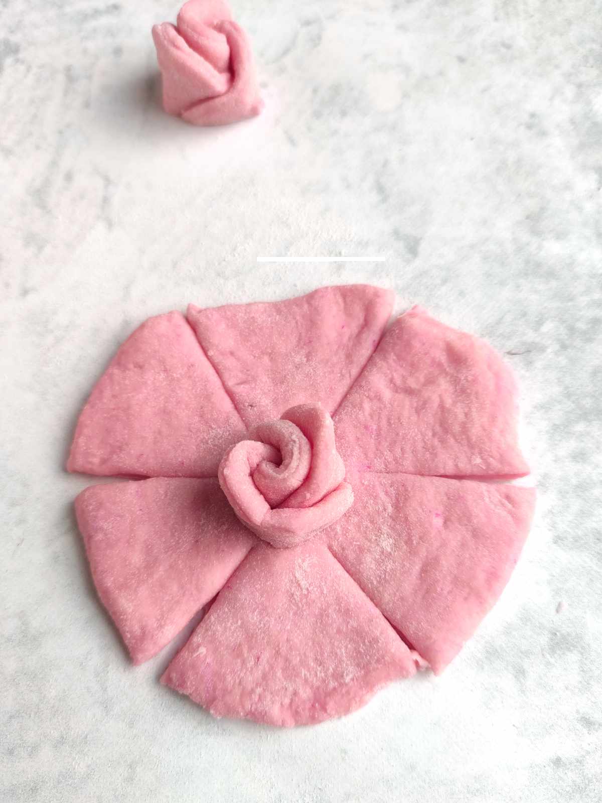 A flat, round piece of pink dough-resembling mantou buns-is cut into six segments with a small rose-shaped dough piece placed at the center. Another rose-shaped dough sits above on a white, lightly floured surface.