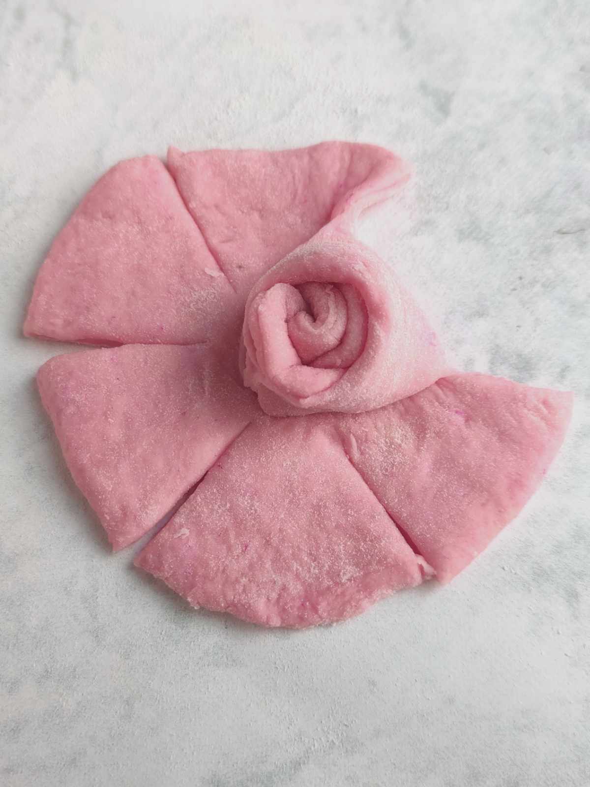 A piece of pink dough, shaped like a flower with a rolled center and five evenly spaced petal-like sections, rests on a lightly floured surface-perfect for making beautiful mantou buns.