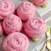 Pink mantou buns shaped like roses are arranged on a light plate, with green dough resembling leaves. Two gold spoons rest nearby on a striped tablecloth. The buns have a soft, swirled texture.