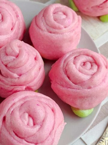 Pink mantou buns shaped like roses are arranged on a light plate, with green dough resembling leaves. Two gold spoons rest nearby on a striped tablecloth. The buns have a soft, swirled texture.