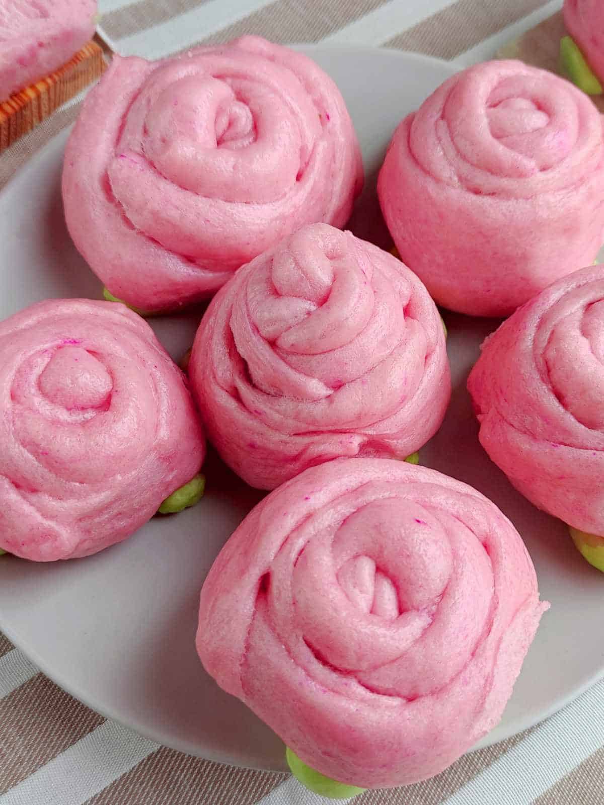 A plate of pink, rose-shaped mantou buns with green bases is arranged neatly on a light-colored plate atop a striped cloth.