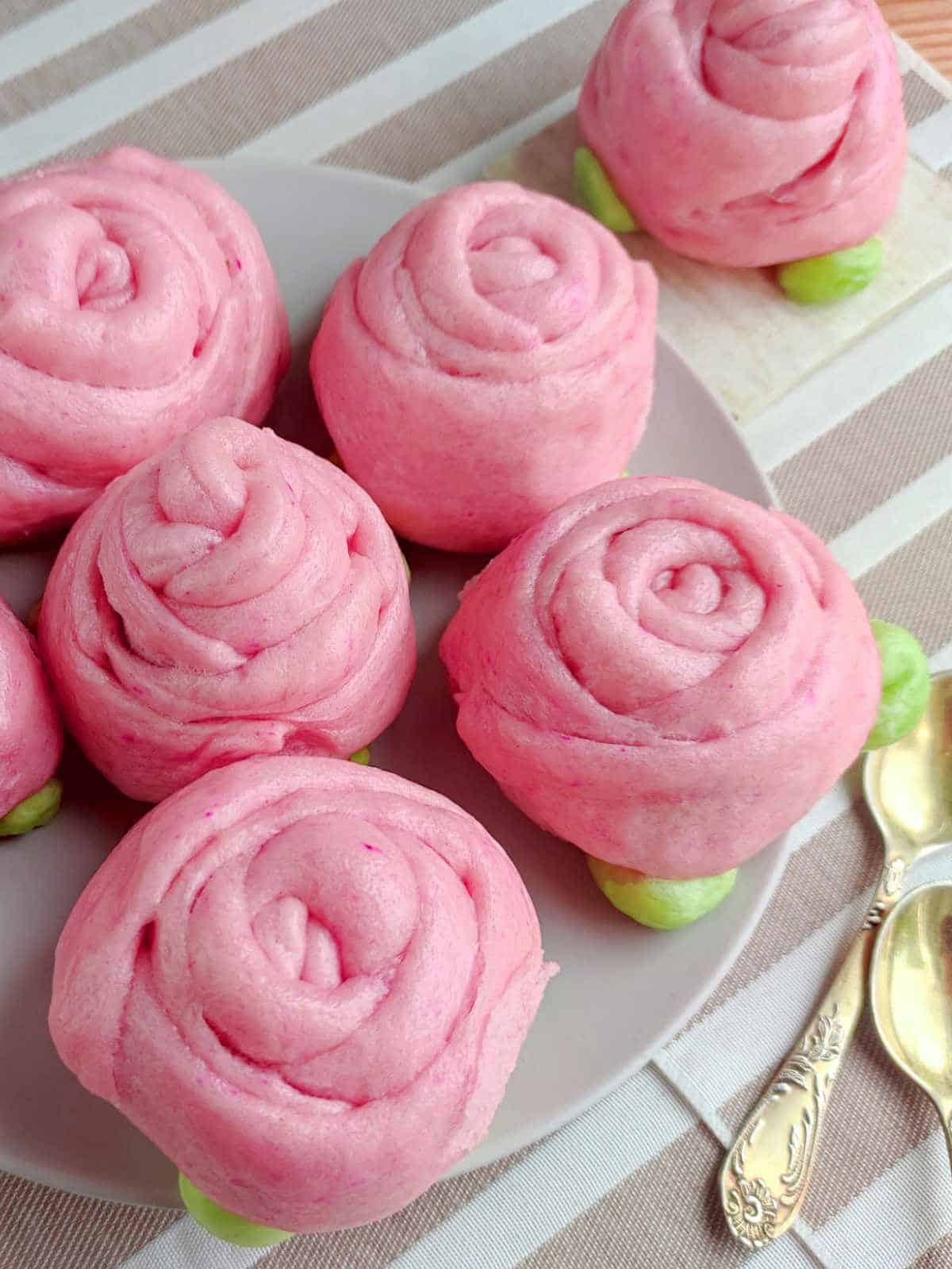 A plate of pink mantou buns shaped like roses with green bases, arranged on a striped cloth next to two ornate gold spoons.