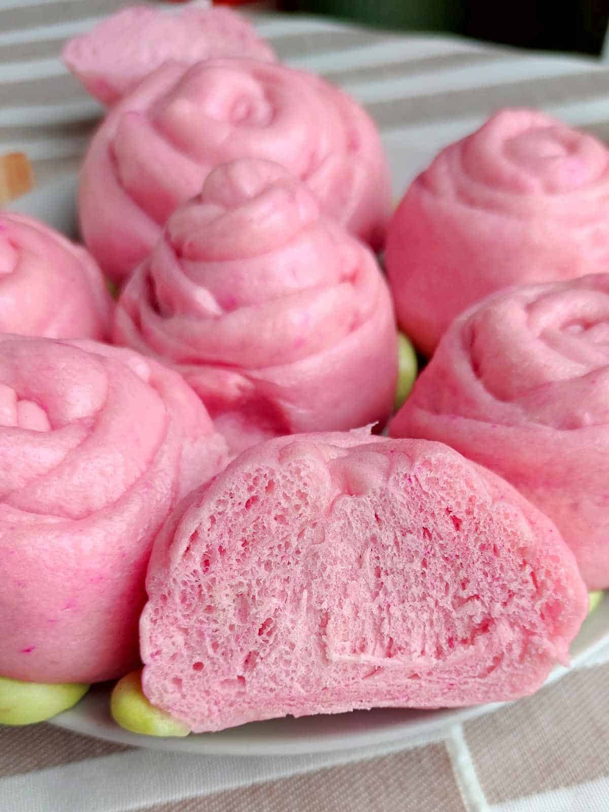 A plate of pink, rose-shaped mantou buns with a soft, fluffy texture. One bun is cut open to reveal the airy interior. The buns are arranged closely together on a white plate.