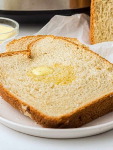 A thick slice of homemade bread with a melting pat of butter on top, served on a white plate. A loaf of bread and a small bowl of butter are in the background.