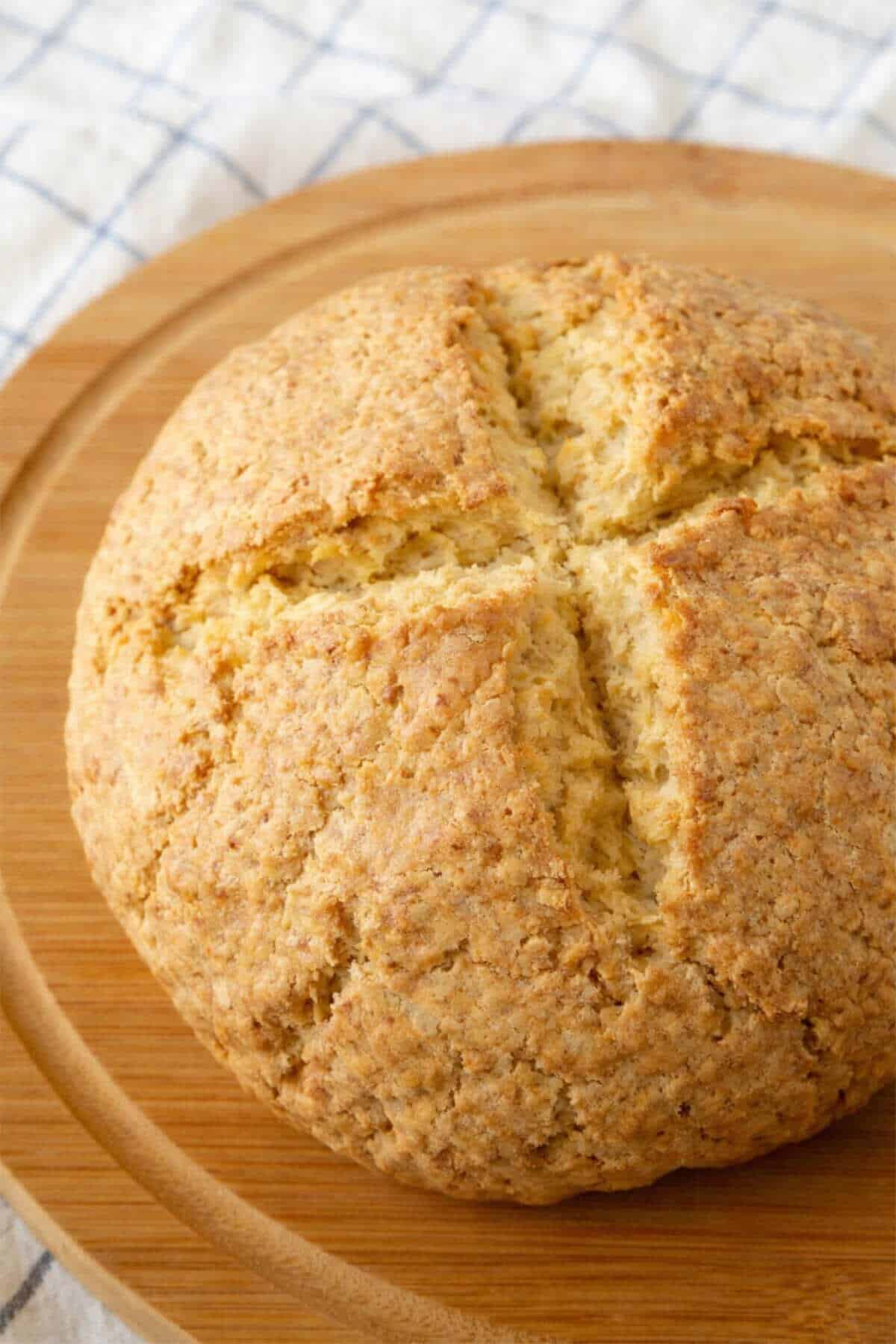 A round loaf of golden-brown gluten free Irish soda bread with a cross cut into the top sits on a wooden board, with a white and blue checkered cloth in the background.