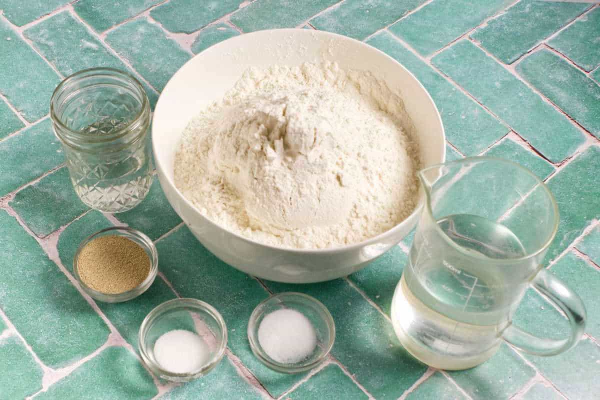 A large white bowl of flour is surrounded by small bowls of yeast, salt, and sugar, plus two clear jars of water, all on a green tiled surface for making a Waterford blaa recipe.