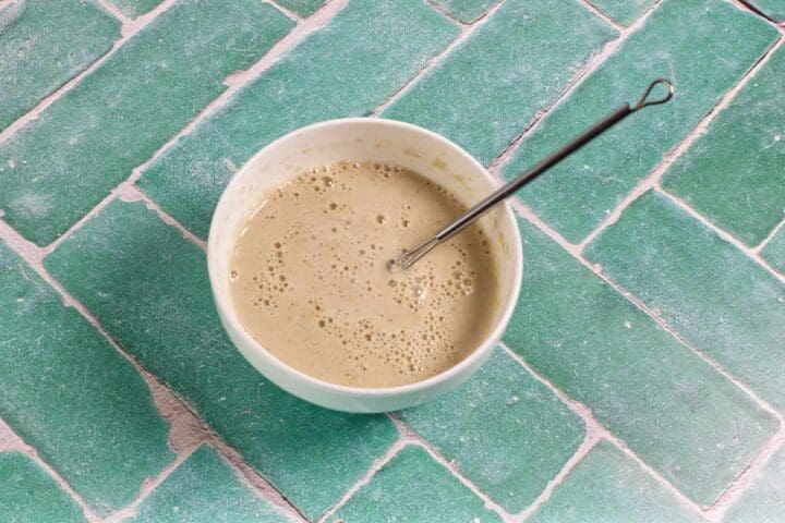 A white bowl filled with a bubbly beige batter or mixture, with a metal whisk resting inside, sits on a green tiled surface.