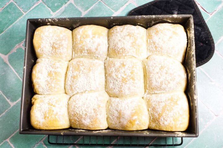 A baking pan filled with twelve pale, flour-dusted Waterford blaa rolls sits on a cooling rack atop green tile. A black oven mitt is partially visible in the top right corner.