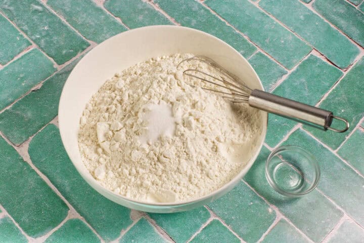 A large white bowl filled with flour and a small pile of salt in the center, with a metal whisk resting on top. A small empty glass sits nearby on a green tiled surface.