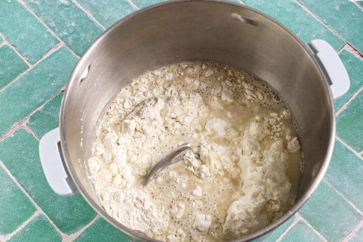 A large metal pot on a green tiled surface contains flour and water being mixed, with a metal spoon partially submerged in the mixture.
