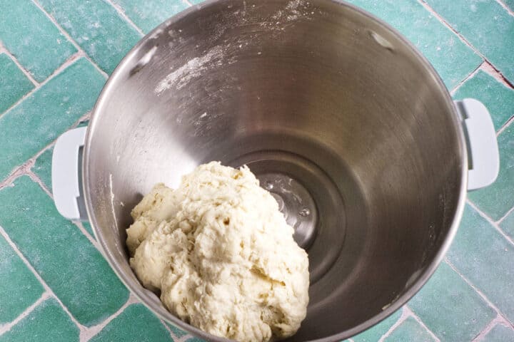 A ball of dough sits in a large metal mixing bowl on a green, patterned surface.