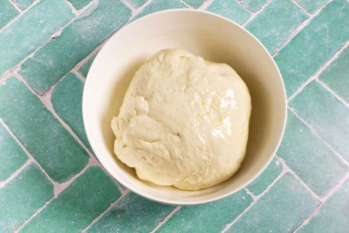 A ball of dough sits in a white bowl on a green tile surface, ready for baking or further preparation.