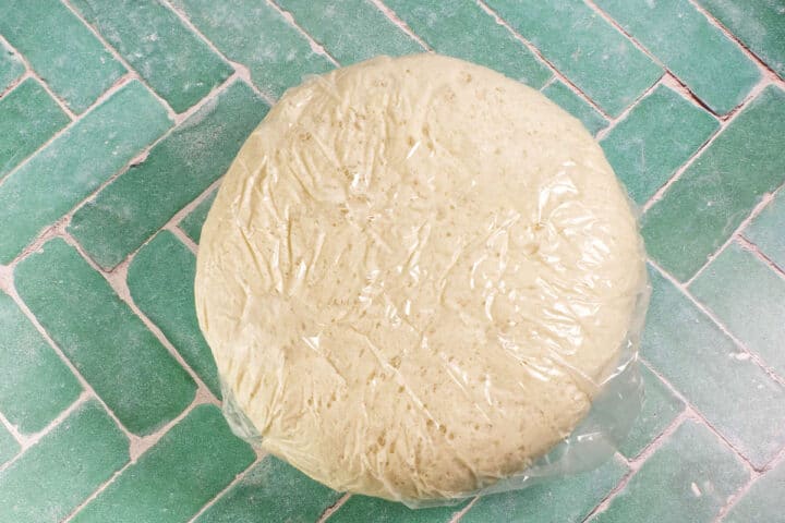A round ball of dough covered with plastic wrap is resting on a green tiled surface.