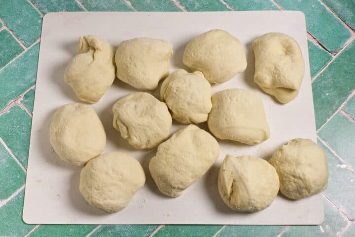 Twelve pieces of raw Waterford blaa dough are arranged on a white cutting board placed on a green tiled surface.