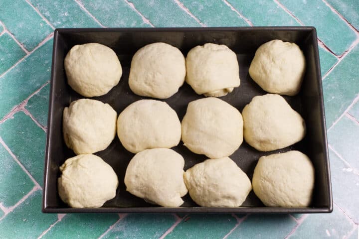 A baking tray with twelve round balls of unbaked Waterford blaa dough arranged in rows on a green tiled surface.