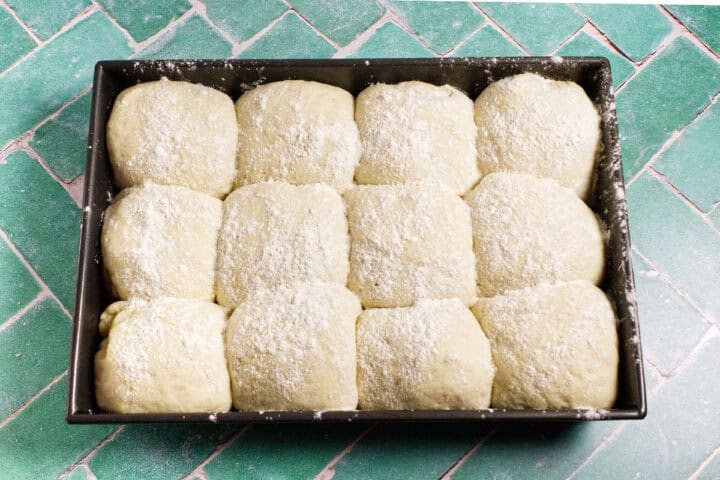 A rectangular baking pan filled with twelve unbaked, floured Waterford blaa rolls arranged in three rows on a green tiled surface.