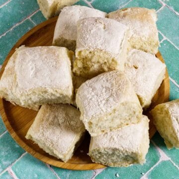 A wooden plate holds several square, fluffy bread rolls dusted with flour, arranged on a green and white tiled surface.
