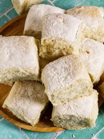A wooden plate holds several square, fluffy bread rolls dusted with flour, arranged on a green and white tiled surface.