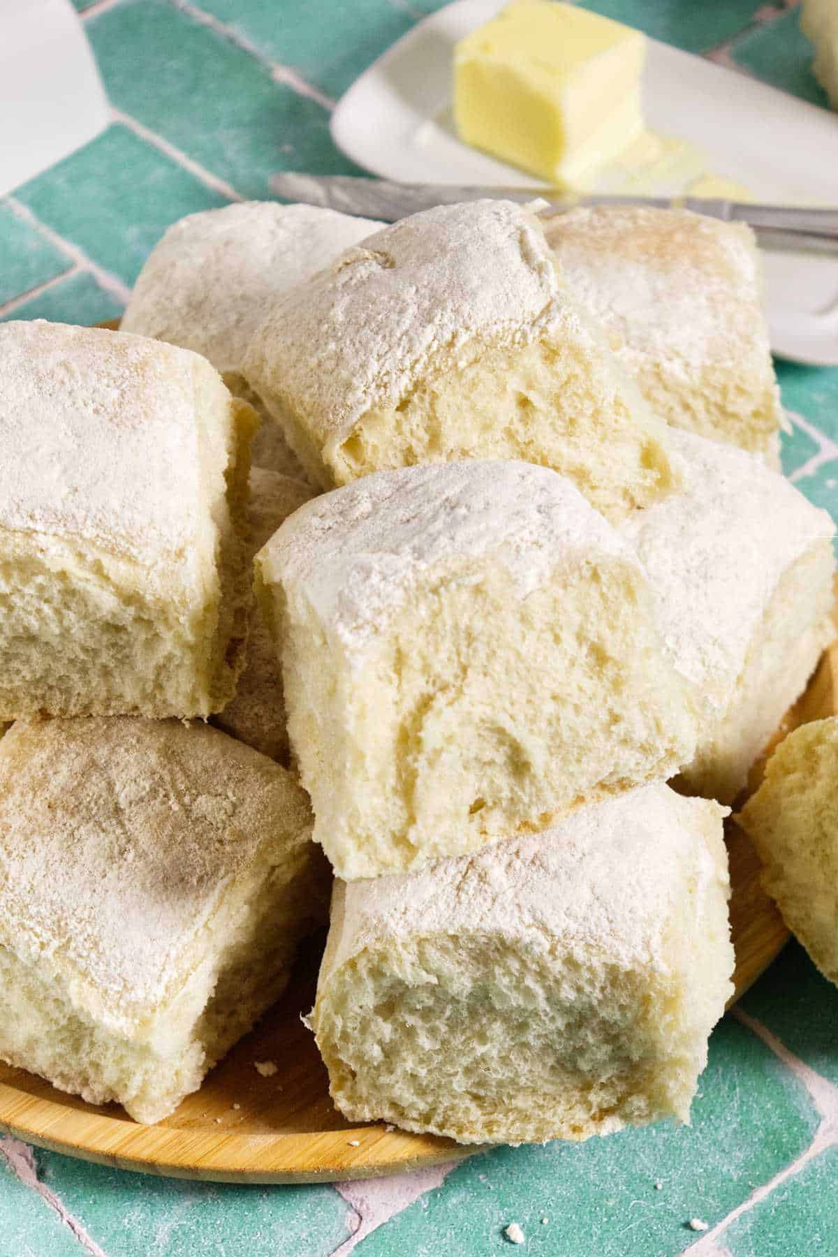 A stack of square Waterford blaa rolls dusted with flour sits on a wooden plate. In the background, a pat of butter rests on a white dish atop a green tiled surface.